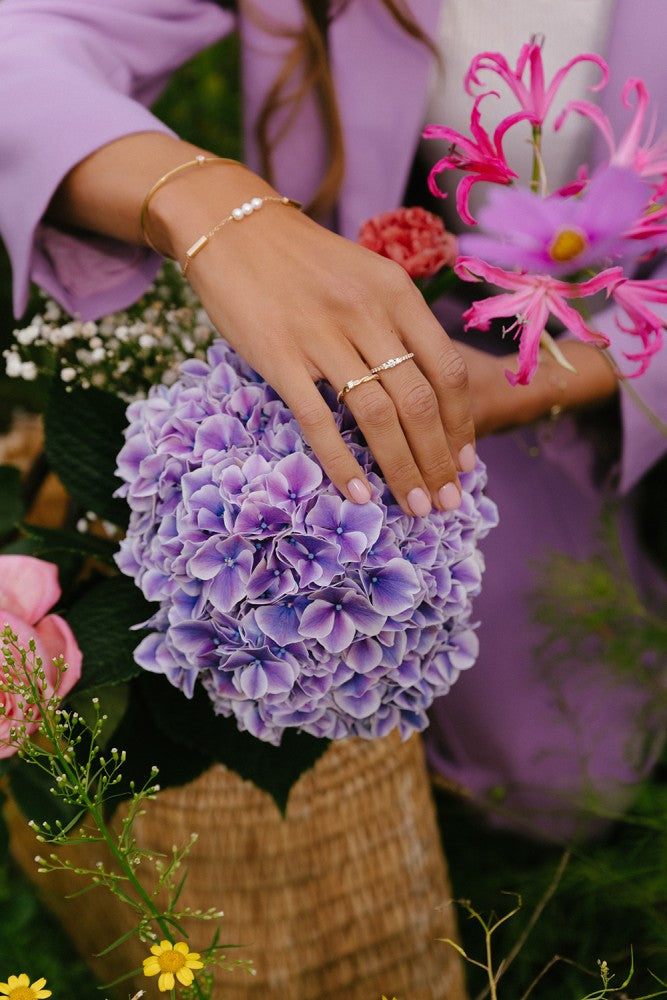 Een vrouwenhand met gouden armbanden en een Excellent Jewelry Geelgouden Driesteens Ring met Extra Diamanten rust op een paarse hortensia. Ze draagt een lavendelblazer en houdt een mand met kleurrijke bloemen vast.