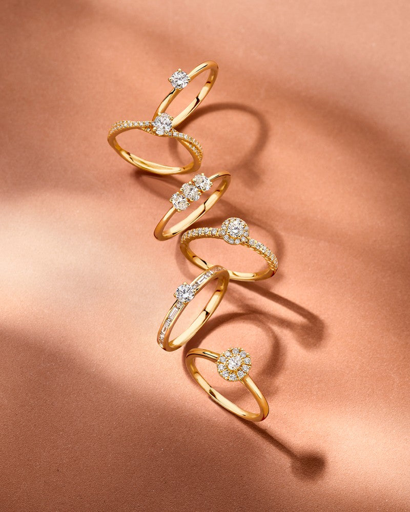 Vijf Elegante Geelgouden Ringen met Ovale Middensteen en Fonkelende Diamanten 0.48 crt. van Excellent Jewelry liggen in een spiraal op een bronzen oppervlak, sfeervol belicht met subtiele schaduwen.
