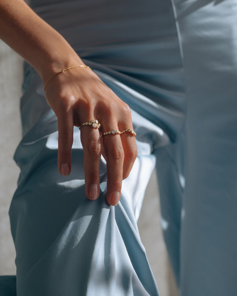 Een close-up van een hand op een gebogen knie, die de Elegante 14k Geelgouden Ring met 0,81ct Diamanten uit de speelse 'Bubbles' Collectie van Excellent Jewelry draagt, plus een gouden armband. Een lichtblauwe satijnen broek en natuurlijk licht creëren zachte schaduwen.