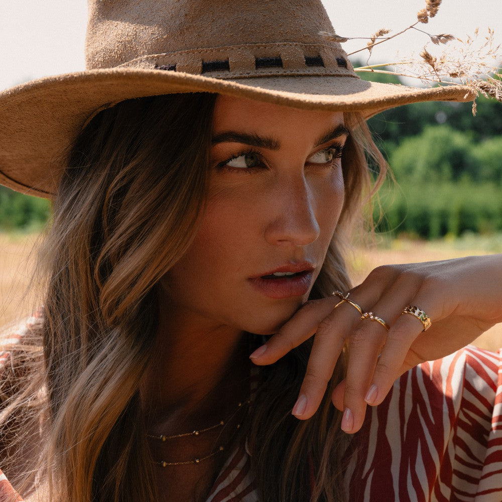 Een vrouw met een bruine hoed en rood gestreept shirt staart peinzend naar buiten, terwijl ze gouden sieraden draagt, waaronder de Elegante Geelgouden Ring met Zirkonia's in Golvend Design - 14 Karaat Goud van Excellent Jewelry.