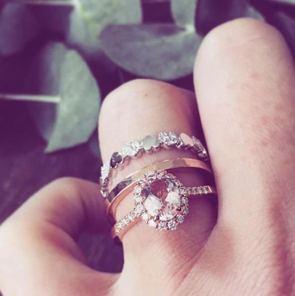 Een close-up van een hand die drie gestapelde ringen draagt: Excellent Jewelry's 14 karaat roségouden ring met 1,28 ct ovale morganiet en 0,27 ct briljanten, een effen gouden band en een zilveren band met kleine steentjes. Vage groene bladeren op de achtergrond.