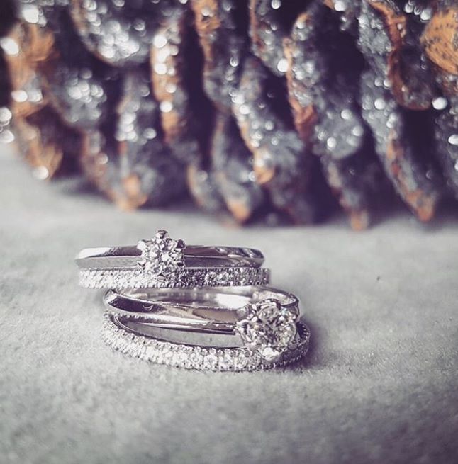 Two Witgouden Ringen met Diamant van Excellent Jewelry, Tijdloze Verfijning in 14 Karaat, liggen op een zachte grijze ondergrond. Op de achtergrond is een wazig, zilverbruin textuurelement zichtbaar.