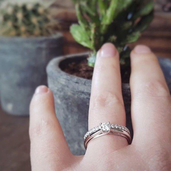 Een close-up van een hand die de Excellent Jewelry Witgouden Ring met Diamant draagt: Tijdloze Verfijning in 14 Karaat, met twee groene planten in potten zacht vervaagd op de achtergrond.
