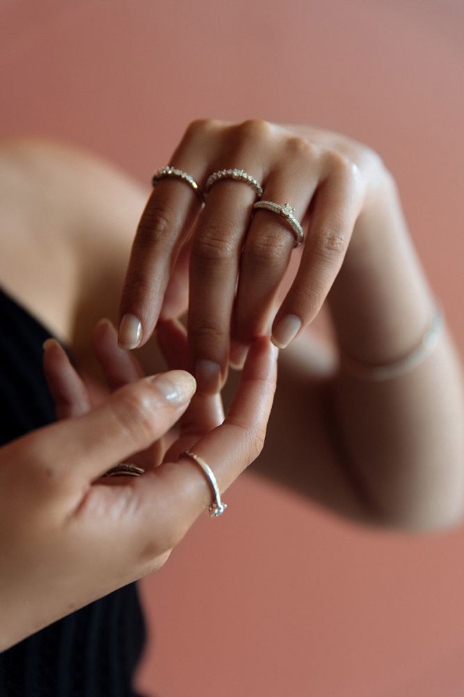 Een close-up van handen die meerdere delicate ringen dragen, waaronder de Excellent Jewelry Stunning White Gold Ring met 0,47 karaat diamanten, op natuurlijke nagels, vingers die elkaar zachtjes aanraken tegen een zacht vervaagde, warmgetinte achtergrond.