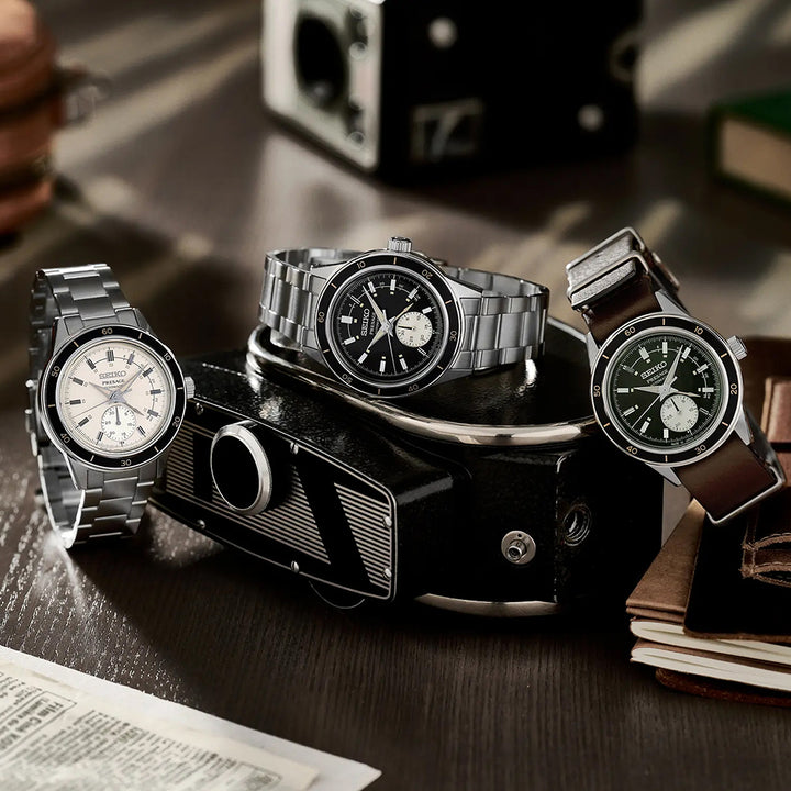 Drie polshorloges, waaronder een Seiko Presage SSA449J1 met een automatisch uurwerk in een roestvrijstalen kast, worden kunstig tentoongesteld op een vintage camera op een houten tafel. De horloges hebben witte, zwarte en groene wijzerplaten met metalen en leren banden. Boeken en papieren liggen nonchalant op de achtergrond.