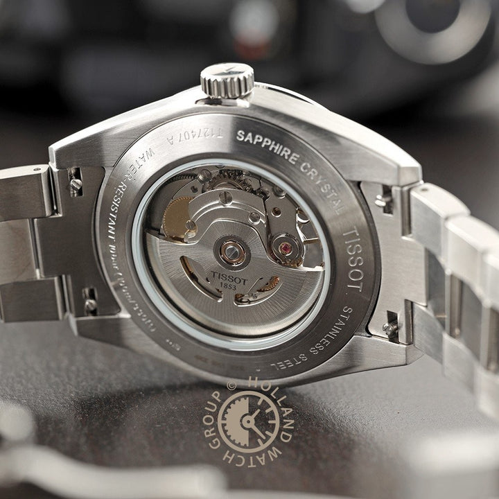 Close-up van de transparante achterkant van de Tissot Gentleman Powermatic 80 Open Heart kast, waarop het automatische uurwerk en de gegraveerde details op de metalen rand te zien zijn, elegant geplaatst op een donker oppervlak.