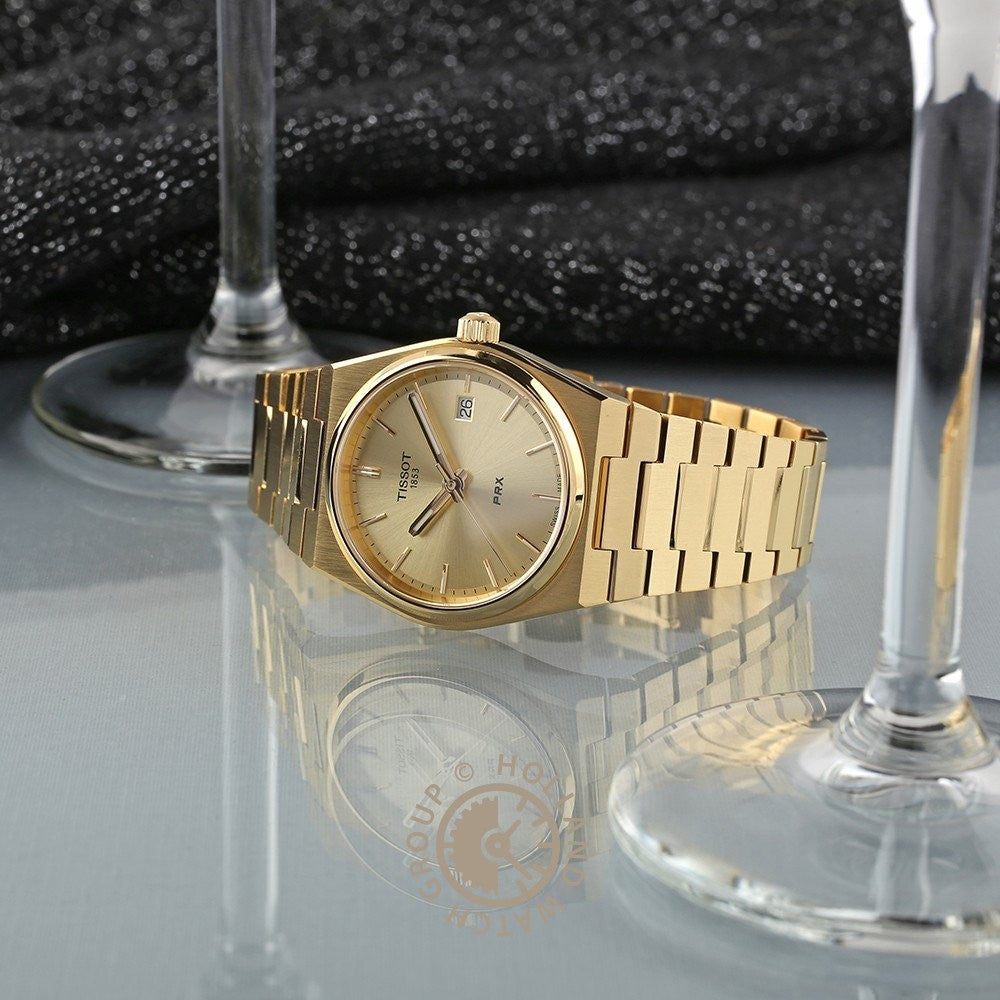 Een Tissot PRX 35mm gouden polshorloge met een metalen band wordt weergegeven op een reflecterend oppervlak tussen twee lege wijnglazen, tegen een donkere, glitterende stof. Dit Tissot horloge is voorzien van Zwitsers kwartsuurwerk en krasbestendig saffierglas.