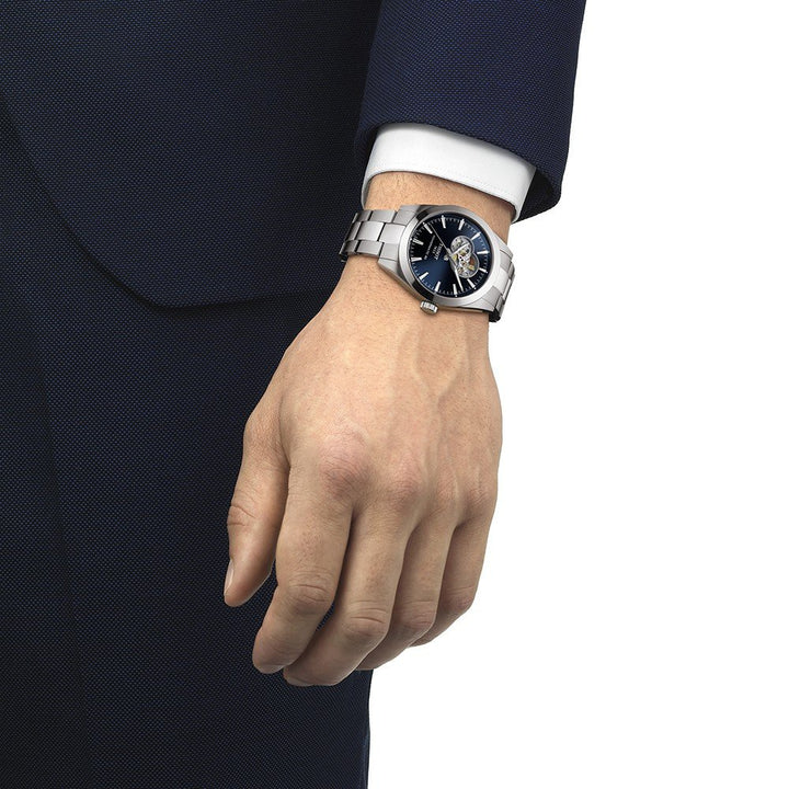 Een persoon in een donkerblauw pak en wit overhemd draagt een Tissot Gentleman Powermatic 80 Open Heart met een blauwe wijzerplaat en zichtbaar Zwitsers automatisch uurwerk om zijn linkerpols.
