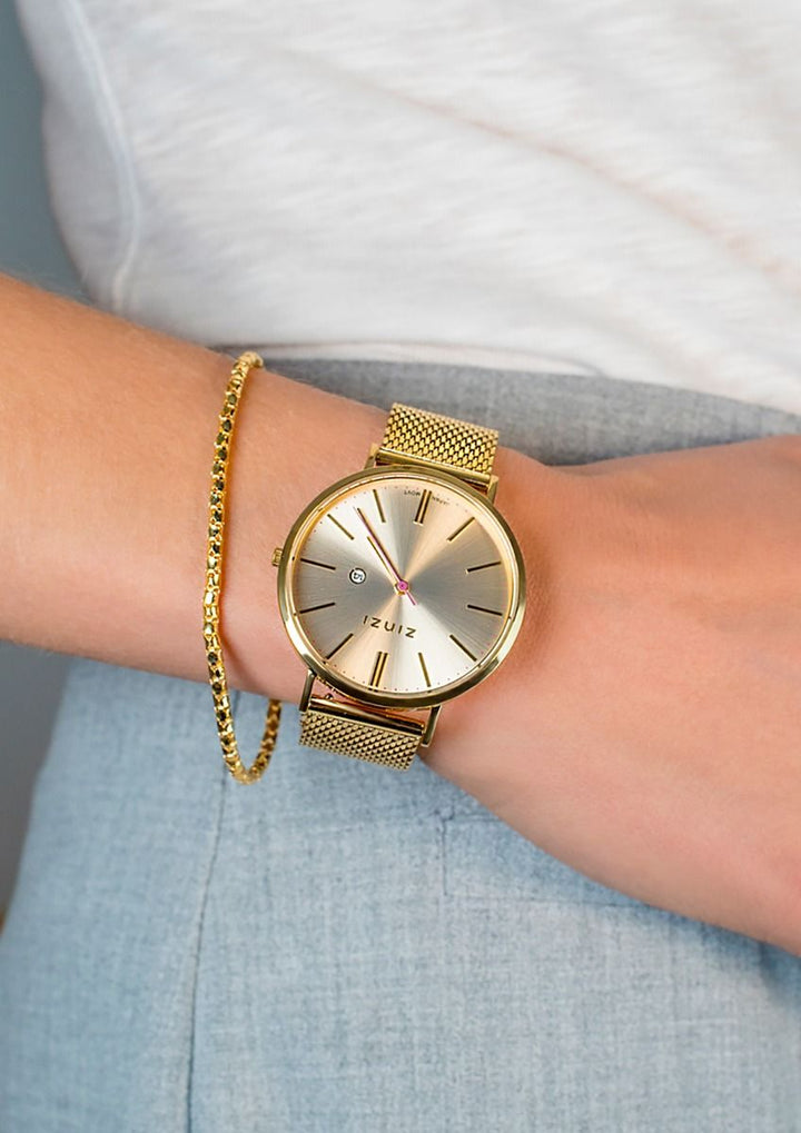 Iemand draagt het Zinzi Goudkleurig Horloge Gold Plated Ziw410 M met een stalen mesh band en goudkleurige wijzerplaat, gestyled met een slanke gouden armband om de linker pols, samen met een lichtblauwe broek en een wit topje.