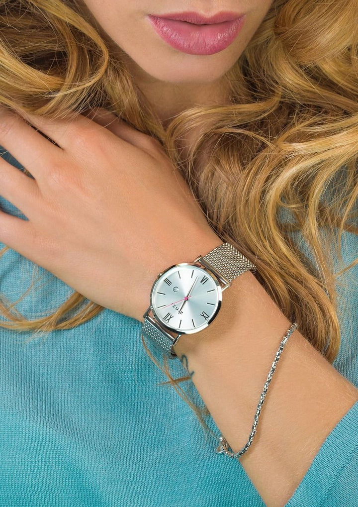 Een vrouw met golvend blond haar draagt een Zinzi Zilverkleurig Horloge Staal ZIW502 M met een witte wijzerplaat en Romeinse cijfers, gekoppeld aan een dunne zilveren armband. Ze is gekleed in een lichtblauw topje, haar hand rust dichtbij haar sleutelbeen.