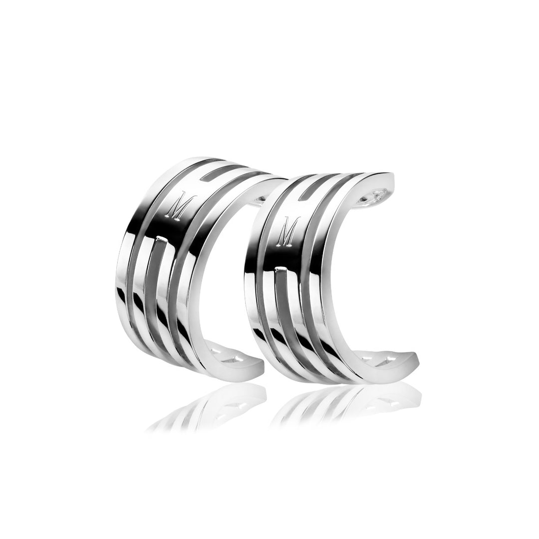 De Zinzi Oorbellen Zilver Mvo1 manchetoorbellen hebben drie parallelle banden met op elke band een gegraveerde M. Ze stralen minimalistische verfijning uit en schitteren elegant op een glanzend wit oppervlak.
